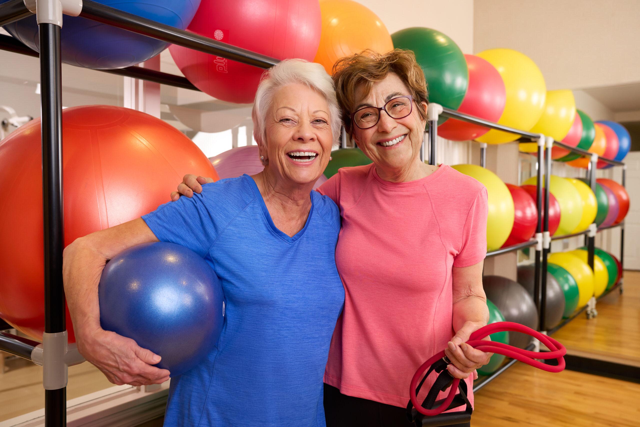 Two women smile for the camera, holding exercise equipment, in front of colorful exercise balls.