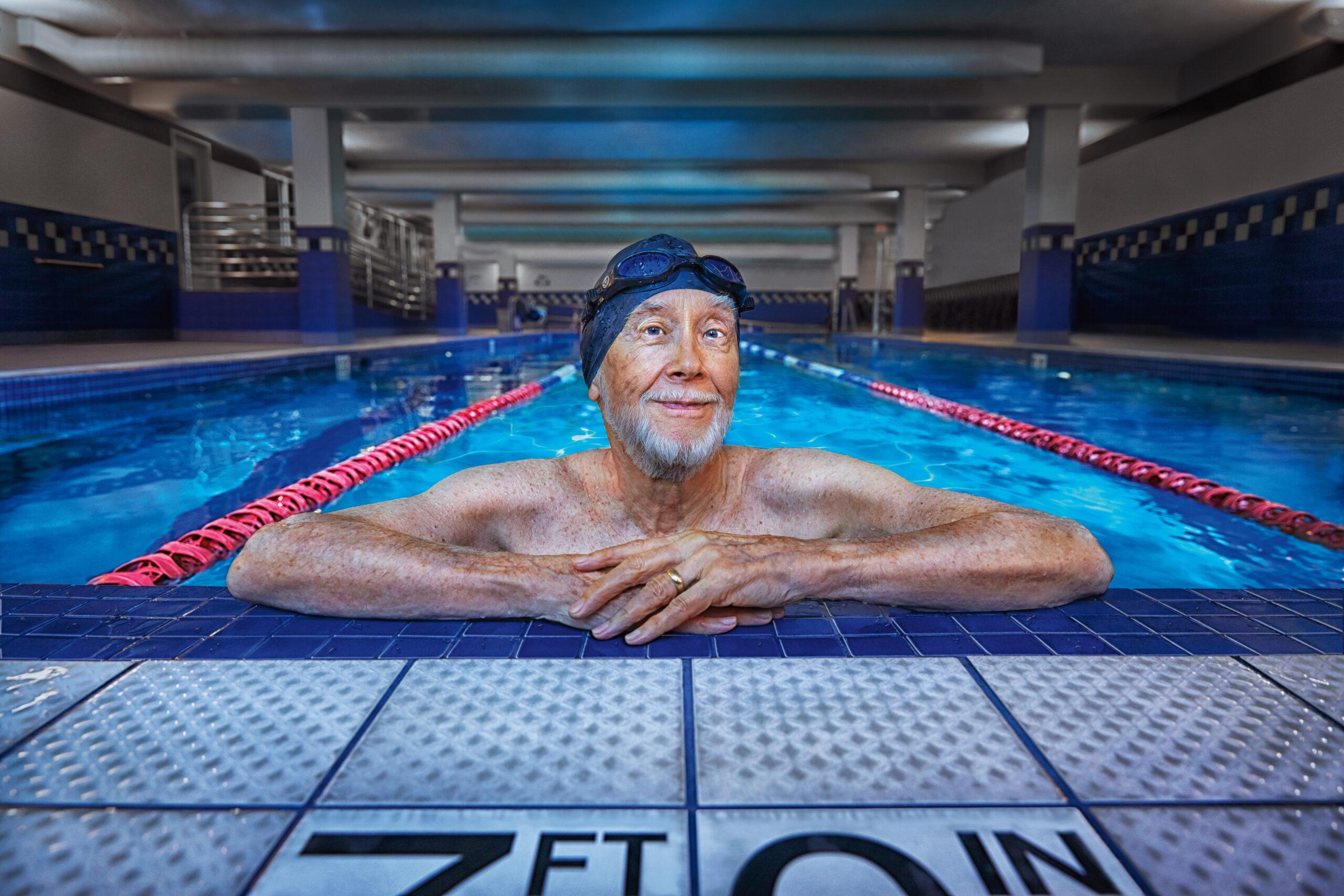 A happy older man wearing a swim cap rests on the side of the pool.