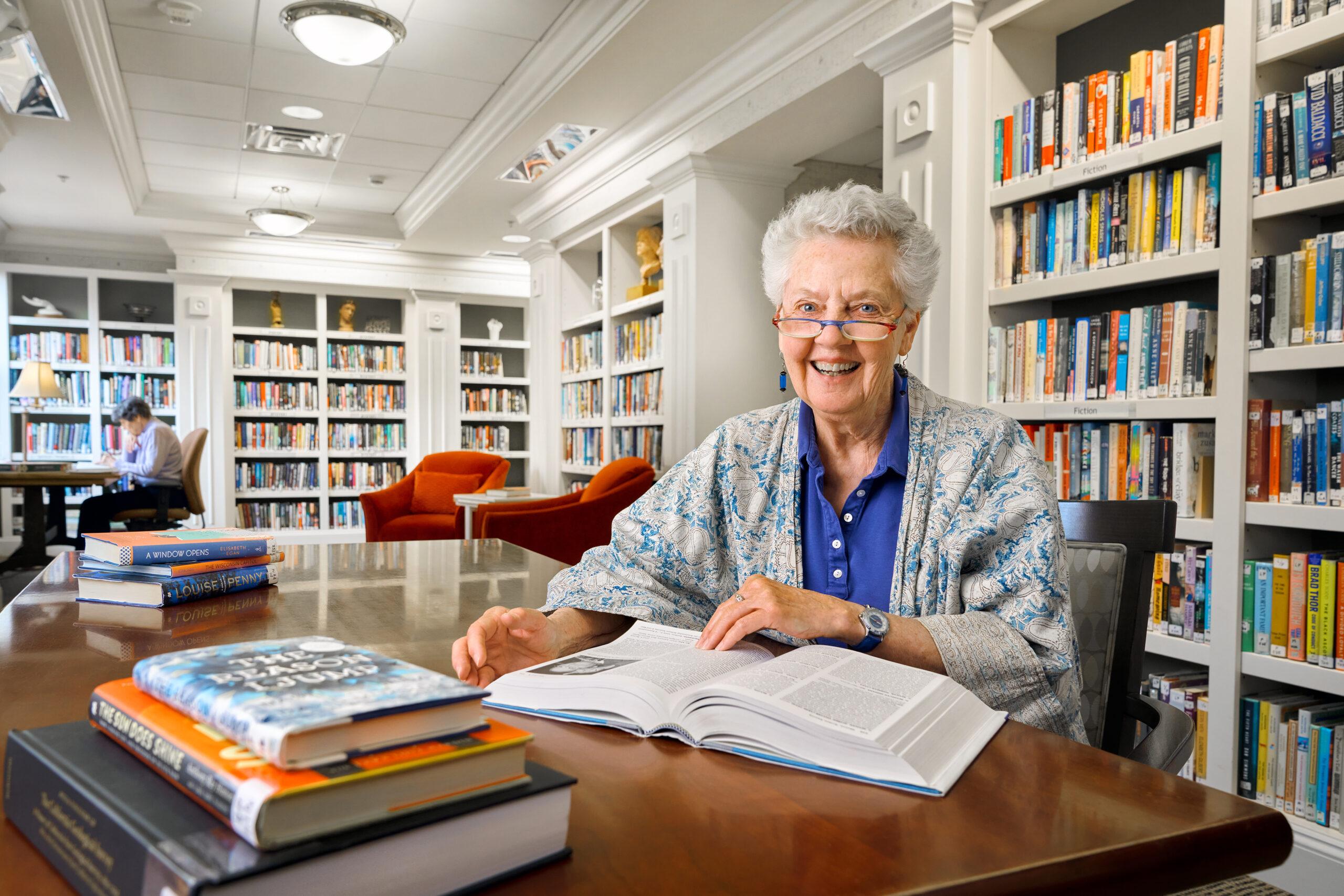 An older woman sitting in a library smiles at the camera as she looks up from her book.