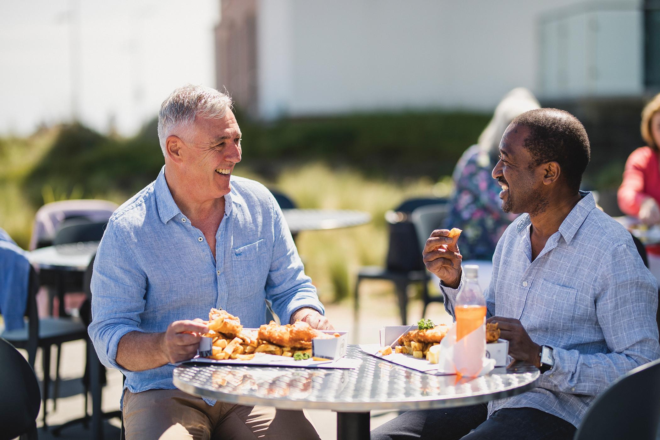 Happy seniors eating fish and chips together in sunny cafe garden.