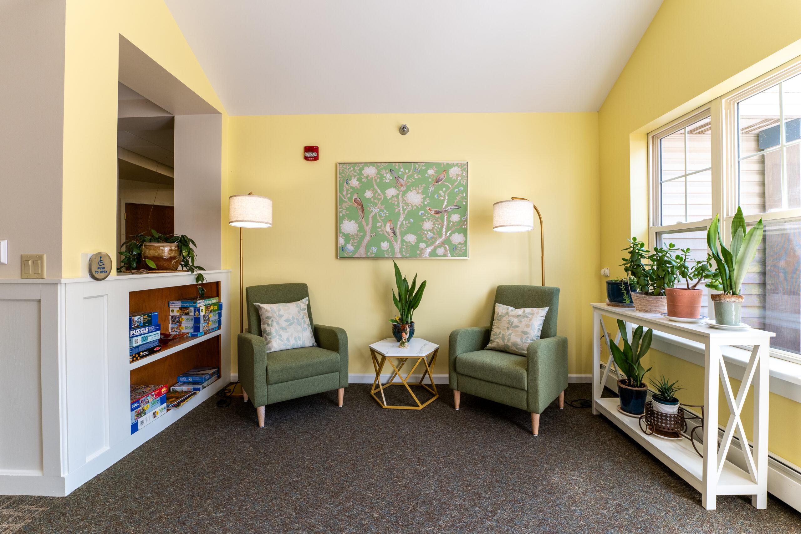 A seating area with yellow wallpaper, green chairs and a shelf of plants.