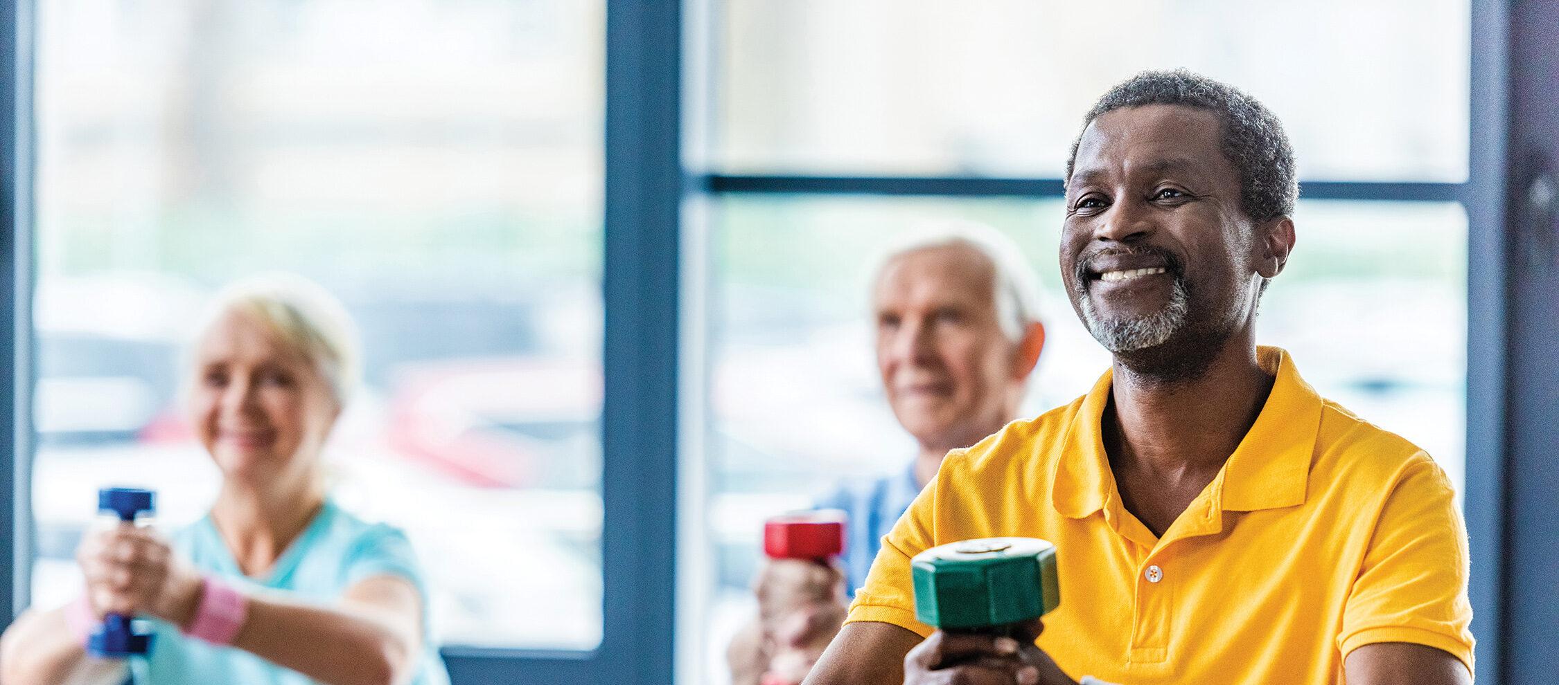 A man smiles as he works out with a dumbbell. Two people are doing a similar workout in the background behind him.